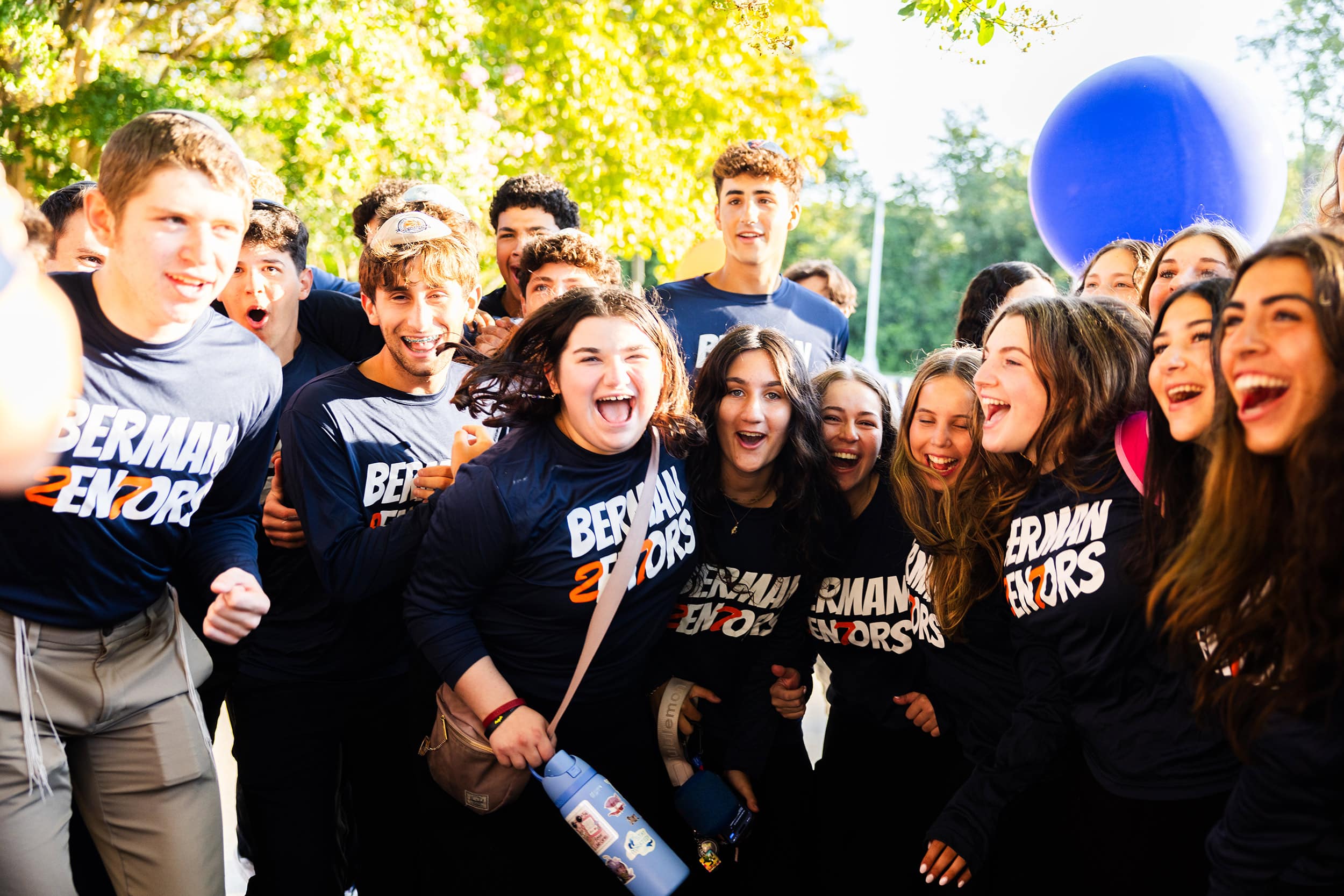 A group of high school students in matching "Berman Seniors" apparel smiling, laughing, and cheering together outdoors on a sunny day, with a large blue balloon visible in the background.